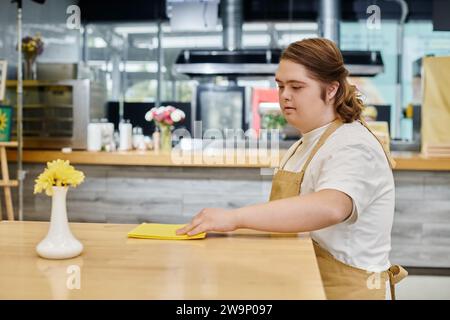 giovane donna con sindrome di down che pulisce il tavolo con lo straccio mentre lavora in una caffetteria moderna, inclusività Foto Stock