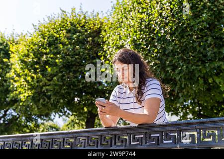 Una gioiosa donna di mezza età con i capelli lunghi e fluenti sta passeggiando in un parco panoramico mentre si trova nel suo smartphone, multitasking e si sta divertendo Foto Stock