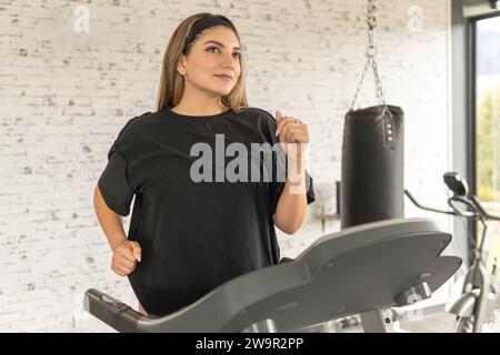 Una giovane donna bionda sorridente che si allena sul tapis roulant in palestra durante la mattina per mantenersi in forma Foto Stock