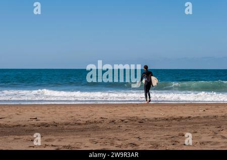 Un surfista maschio sta per entrare nell'oceano con la sua tavola da surf e la muta. Giornata soleggiata e ventosa nella splendida spiaggia di Sopelana a nord di Bilbao Foto Stock