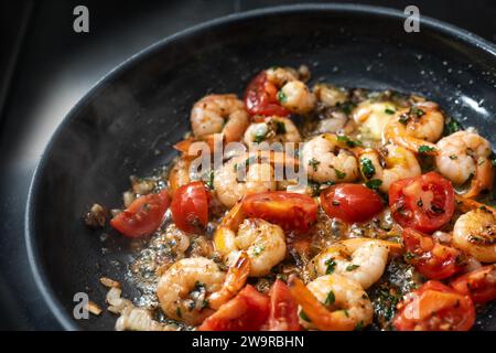 I gamberetti con coda vengono saltati con pomodori, erbe e aglio in olio d'oliva in una padella nera, cucinando un pasto mediterraneo a base di pesce, focu selezionato Foto Stock