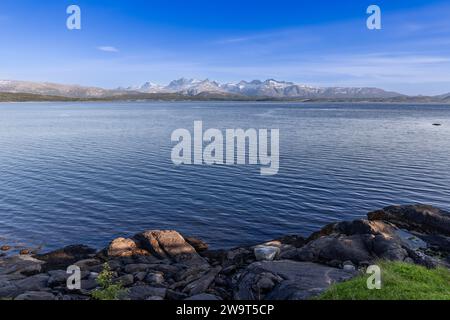 Vista estiva della tranquilla Saltfjorden con montagne innevate, grandi massi in primo piano, vicino a Bodo, Isole Lofoten, Norvegia Foto Stock