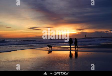 Gli amanti delle passeggiate con i cani fanno una passeggiata all'alba lungo la spiaggia di Tynemouth, Tyne and Wear, sulla costa nord-orientale dell'Inghilterra. Durante l'ultimo fine settimana dell'anno sono in arrivo raffiche di neve come parte del brace britannico, sulle orme di pochi giorni ventosi dovuti a Storm Gerrit. Data immagine: Sabato 30 dicembre 2023. Foto Stock