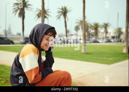 Allegro adolescente latinoamericano, hipster boy parla al cellulare, seduto su un prato verde nel parco cittadino. Persone. Tecnologia wireless e Internet Foto Stock