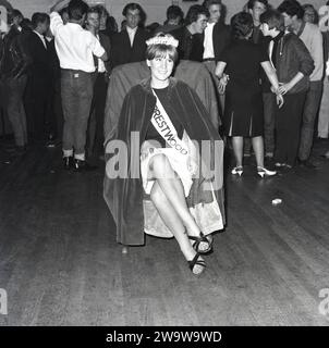 1964, storica, bella regina.... Al centro di un municipio con altri giovani presenti, una giovane donna, con corona, fascia e mantello seduto su una sedia dopo essere stata incoronata come "Miss Prestwood". L'antico villaggio di Prestwood nelle Chilterns, vicino a Great Missenden nel Buckinghamshire, Inghilterra, risale all'epoca anglosassone. Foto Stock