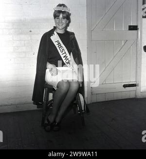 1964, storica, una giovane donna seduta su una sedia di legno in un angolo di una sala del villaggio con corona, fascia e mantello dopo un concorso di bellezza e incoronata Miss Prestwood. L'antico villaggio di Prestwood nelle Chilterns, vicino a Great Missenden nel Buckinghamshire, Inghilterra, risale all'epoca anglosassone. Foto Stock