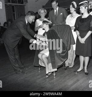 1964, storica, bella regina.... Nel mezzo di un municipio con giudici in presenza, una giovane donna, con corona, fascia e mantello seduto su una sedia che riceve un regalo dopo essere stata incoronata come 'Miss Prestwood' dell'anno. L'antico villaggio di Prestwood nelle Chilterns, vicino a Great Missenden nel Buckinghamshire, Inghilterra, risale all'epoca anglosassone. Foto Stock