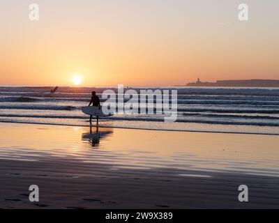 Il surfista esce dal mare mentre un altro entra su una spiaggia sabbiosa al tramonto con l'isola alle spalle nella città di Essaouira, in Marocco. 30 dicembre 2023 Foto Stock