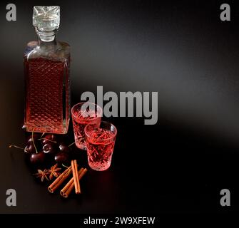 Liquore alla ciliegia con spezie, alcol forte in un decanter e due bicchieri su sfondo nero, bacche mature e cannella con anice. Primo piano. Foto Stock