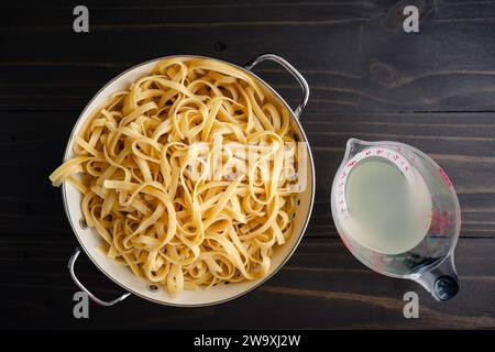 Fettuccine cotte in un colander con una tazza di acqua di pasta: Spaghetti bolliti e sgocciolati in un colander con un dosatore di acqua di cottura riservata Foto Stock