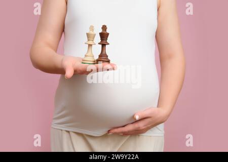 Donna incinta che tiene in mano pezzi di scacchi, sfondo rosa studio Foto Stock