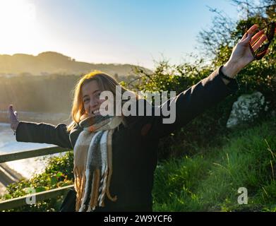 ritratto di una bionda sorridente che indossa una giacca di lana e una sciarpa marrone a scacchi. Sullo sfondo dell'immagine è possibile vedere un paesaggio costiero wi Foto Stock