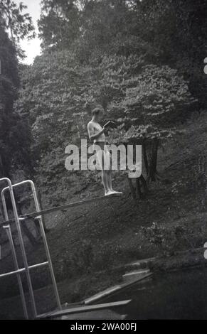 Anni '1950, storico, un nuotatore maschile in piedi su un trampolino, leggendo un libro, in una piscina boschiva all'aperto, Inghilterra, Regno Unito. Foto Stock