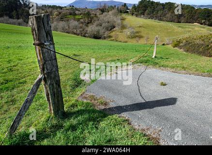 primo piano di un vecchio palo in legno contrassegnato da cui è appeso un cavo in acciaio arrugginito che corre sullo sfondo a un altro palo, isolato che blocca la strada Foto Stock