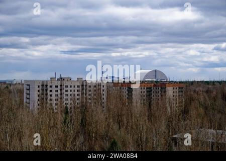 Un vecchio edificio a più piani sorge in mezzo alla vegetazione ricoperta, con una struttura a cupola in lontananza. Foto Stock