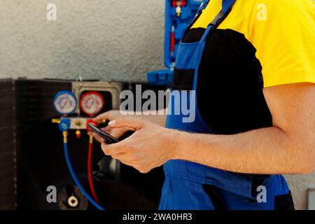 Elettricista capace che ordina nuovi componenti per condensatore difettoso dopo aver terminato la manutenzione. Primo piano del wireman meticoloso che cerca online sul telefono parti di ricambio per sistemi hvac esterni Foto Stock