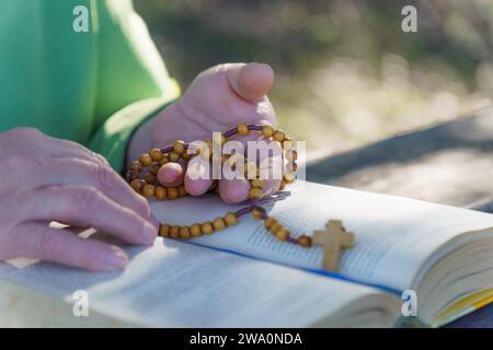 Le mani di una donna che prega il rosario sul campo sopra una bibbia aperta su un tavolo di legno Foto Stock