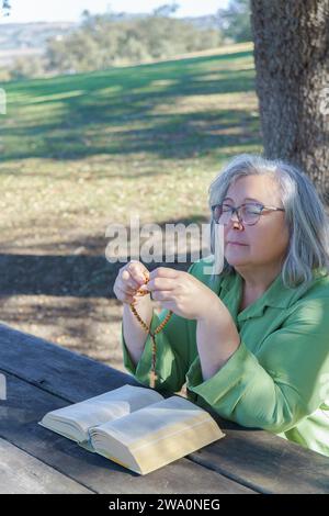 Donna dai capelli bianchi più anziana con perline di rosario in mano e una bibbia che prega con gli occhi chiusi seduto a un tavolo di legno nella foresta Foto Stock