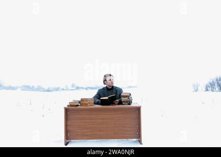 Un giovane e bello uomo intelligente con una giacca siede a un tavolo in un campo e legge libri antichi in inverno Foto Stock