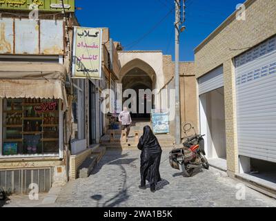 La donna musulmana vestita con il tradizionale chador nero cammina in uno stretto ingresso al Gran Bazar di Kashan, Iran. Foto Stock