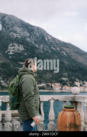 Il viaggiatore con un thermos e uno zaino si trova sulla riva del mare alla ringhiera e guarda le montagne. Vista laterale Foto Stock
