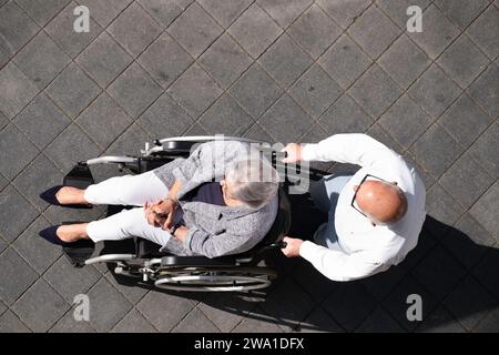 Coppia più anziana vista dall'alto in estate. L'uomo spinge sua moglie su una sedia a rotelle lungo il marciapiede. Copia spazio Foto Stock