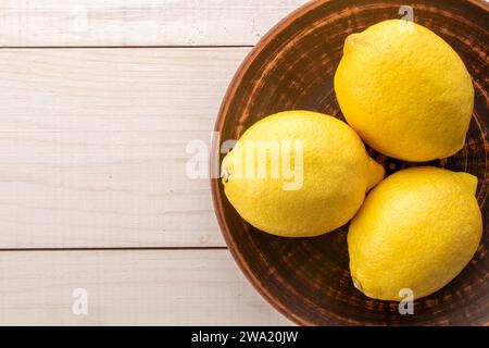 Tre limoni maturi gialli su un piatto di argilla su un tavolo di legno, vista ravvicinata dall'alto. Foto Stock