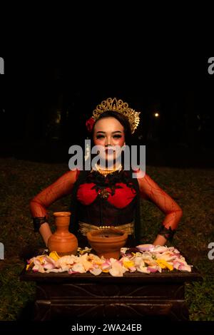 Ripresa ravvicinata di una ragazza asiatica in abiti rossi seduta sulle ginocchia di fronte a un'offerta di fiori di notte Foto Stock