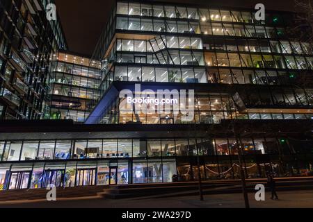 Vista serale sulla sede centrale del Booking.com ad Amsterdam, Paesi Bassi, con finestre illuminate nella facciata di vetro Foto Stock