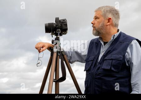 Fotografo al lavoro, capelli grigi e barba folta, 56 anni, gilet fotografico blu scuro, occhiali in mano, fotocamera reflex da 35 mm su treppiede, cielo grigio coperto Foto Stock