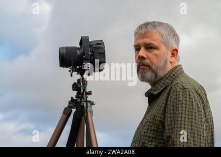 Fotografo al lavoro, capelli grigi e barba folta, 56 anni, look premuroso, camicia a quadri verde oliva, fotocamera reflex da 35 mm su treppiede, cielo grigio Foto Stock
