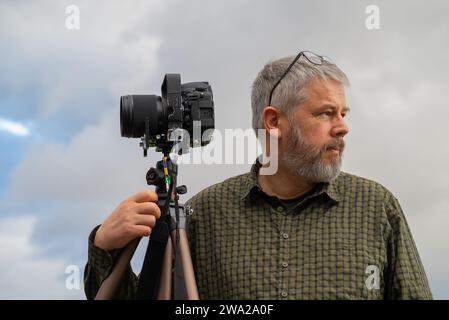 Fotografo al lavoro, capelli grigi e barba piena, 56 anni, occhiali sollevati, guardando in direzione opposta, camicia a quadri verde oliva, reflex da 35 mm Foto Stock