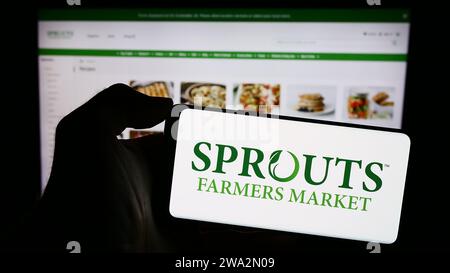 Persona in possesso di un cellulare con il logo della società di vendita al dettaglio statunitense sprouts Farmers Market Inc. Davanti alla pagina Web aziendale. Concentrarsi sul display del telefono. Foto Stock