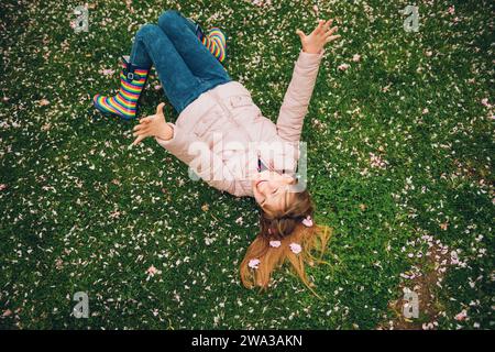 Divertente bambina sdraiata su un'erba verde brillante, giocando con petali di fiori morbidi nel parco primaverile, indossando giacca e stivali da pioggia, moda stagionale per bambini Foto Stock