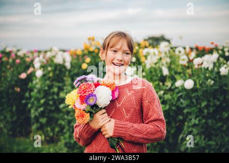 Ritratto all'aperto di una graziosa bambina con un bouquet colorato di fiori dahlia e un pullover alla moda Foto Stock