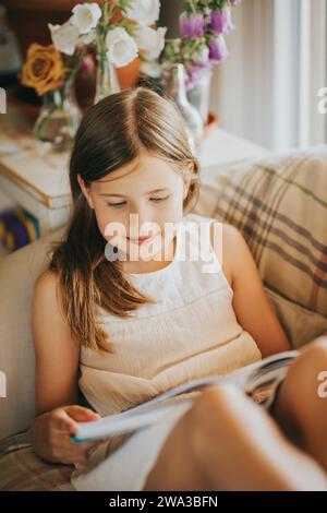 Adorabile bambina che riposa in una comoda poltrona e legge un libro di storia Foto Stock