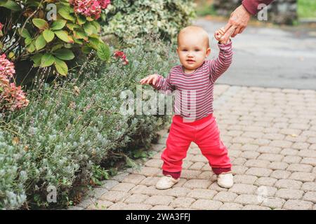 Adorabile bambina di 9-12 mesi che gioca all'aperto, indossa un corpo rosa e jogger, tenendo la mano della madre. I primi passi del bambino, i bambini imparano a camminare Foto Stock