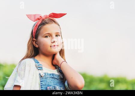 Ritratto all'aperto di una graziosa bambina che indossa tute in denim e fascia rossa vintage, mani nelle tasche Foto Stock
