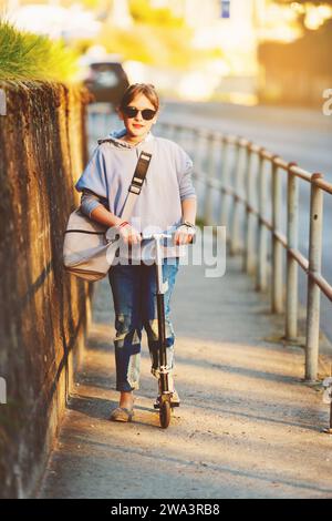 Felice studentessa di 9 anni con zaino per le strade della città al tramonto in scooter, indossando occhiali da sole Foto Stock
