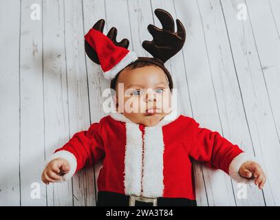 Ritratto natalizio di un adorabile bambino sdraiato su sfondo bianco, indossando l'abito di Babbo Natale e le orecchie delle renne, vista dall'alto Foto Stock