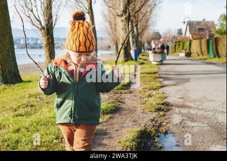 Grazioso bambino di 3 anni che gioca all'aperto nelle soleggiate prime giornate di primavera, indossando una giacca verde e un cappello arancione Foto Stock