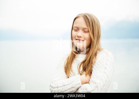 Graziosa bambina di 8 anni che gioca vicino al lago in una giornata molto ventosa, indossa un caldo pullover a maglia bianca, occhi chiusi, braccia incrociate, immagine chiave Foto Stock