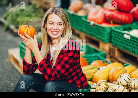 Ritratto all'aperto di una donna bionda felice che tiene in mano una piccola zucca e indossa una camicia a quadri rossa Foto Stock