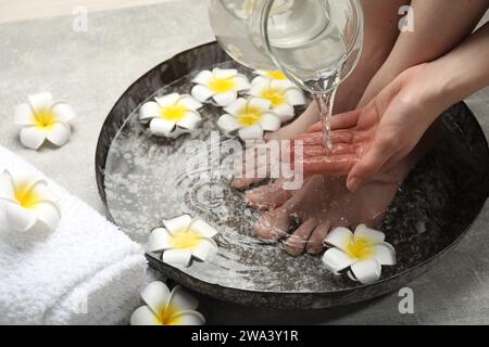 Donna che versa acqua sulla mano mentre ammorbidisce i piedi in un recipiente grigio chiaro, primo piano. Trattamento spa Foto Stock