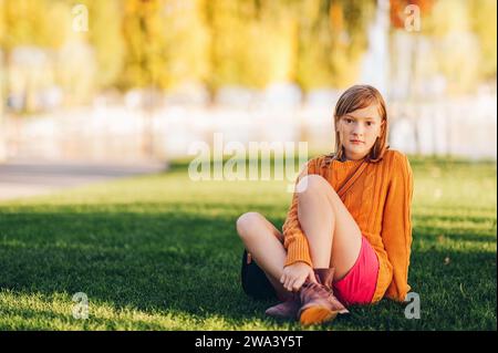 Ritratto all'aperto di una dolce bambina preadolescente, seduta su un prato verde brillante, con maglione arancione e pantaloncini rossi Foto Stock