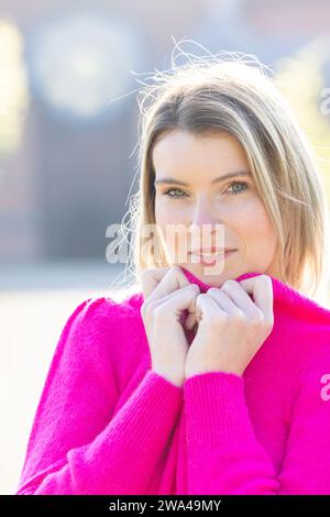 Questo ritratto mostra una giovane donna caucasica con uno sguardo accattivante e un leggero sorriso, che irradia un senso di dolce fascino. Indossa un fantastico maglione fucsia che si stringe al collo, per offrire stile e comfort sullo sfondo morbido. Il sottile gioco di luci e ombre sul suo volto accentua le sue caratteristiche, mentre lo sfondo suggerisce un ambiente di parco urbano. Dolce Allure nel calore della fucsia. Foto di alta qualità Foto Stock