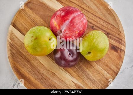 Una varieta' di prugne fresche sistemate su un supporto rustico di legno su un pulito ed elegante sfondo di marmo bianco. La frutta colorata offre un'abbondanza Foto Stock