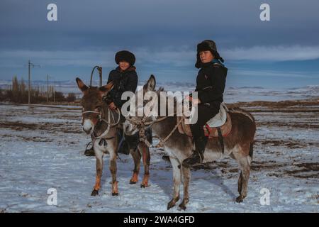 TAU SAMALY, KAZAKISTAN - 23 DICEMBRE 2023: Due amici kazaki ragazzi che cavalcano un asino in inverno in un campo vicino a Tau Samaly in Kazakistan Foto Stock