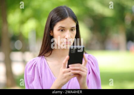 Donna perplessa che controlla strane notizie al telefono in un parco Foto Stock