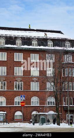 Un team di lavoratori che pulisce la neve dal tetto di un edificio residenziale a più piani dopo la nevicata. Helsinki, Finlandia. 2023. Foto del telefono. Foto Stock